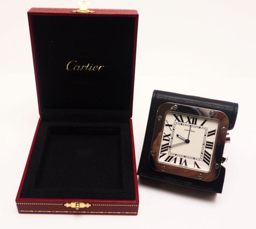 Lote 4022 - CARTIER, RELÓGIO DESPERTADOR - relógio despertador de viagme da marca Cartier, com caixa quadrada em aço prateado, mostrador branco com numeração romana a preto, ponteiros luminiscentes, com suporte em pele preta, em esti«ojo original. Preço 