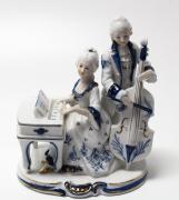 Lote 23 - CENÁRIO MUSICAL EM PORCELANA - Representando casal de músicos tocando Piano e Violão. Decoração em tons de azul com elementos pintados a ouro. Dim: 17.5x14x10cm. Nota: Muito bom estado geral
