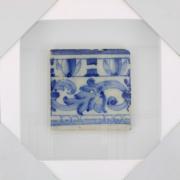 Lote 57 - AZULEJO, SÉC. XVIII - Azulejo antigo aplicado em moldura vitrine (branca). Decoração em tons de com motivos vegetalistas e geométricos. Dim: 14x14 cm (azulejo), 29,5x29,5 cm (moldura). Nota: sinais de manuseamento
