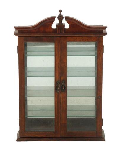 Lote 4 - VITRINE EM MADEIRA ESCURECIDA - Com 2 portas em vidro e interior com 4 prateleiras em vidro translúcido e fundo espelhado. Corpo encimado por pináculo entalhado. Ferragens em metal bronzeado. Dim: 67x44x11 cm