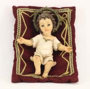 Lote 189 - MENINO JESUS - Figura de Menino Jesus deitado em resina com decoração policromada e dourada repousando em almofada bordeaux com galões dourados. Dim: 25 cm (Menino)