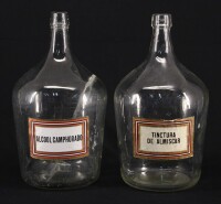 Lote 96 - GARRAFÕES DE VIDRO - Par de garrafões de 5 litros em vidro translúcido, com etiquetas inscritas "Alcool Camphorado" e "Tintura de Almiscar", vazios. Dim: 35 cm (altura)