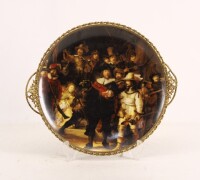 Lote 109 - PRATO DECORATIVO EM PORCELANA - Decoração policromada com detalhe da pintura "A Ronda da Noite" de Rembrandt. Aro e pegas em metal dourado. Dim: 30x34 cm